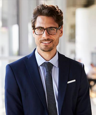 Momento Invertir mis ahorros - Joven hombre de negocios con traje azul, corbata y camisa azul clarito y gafas, sonriendo en la oficina